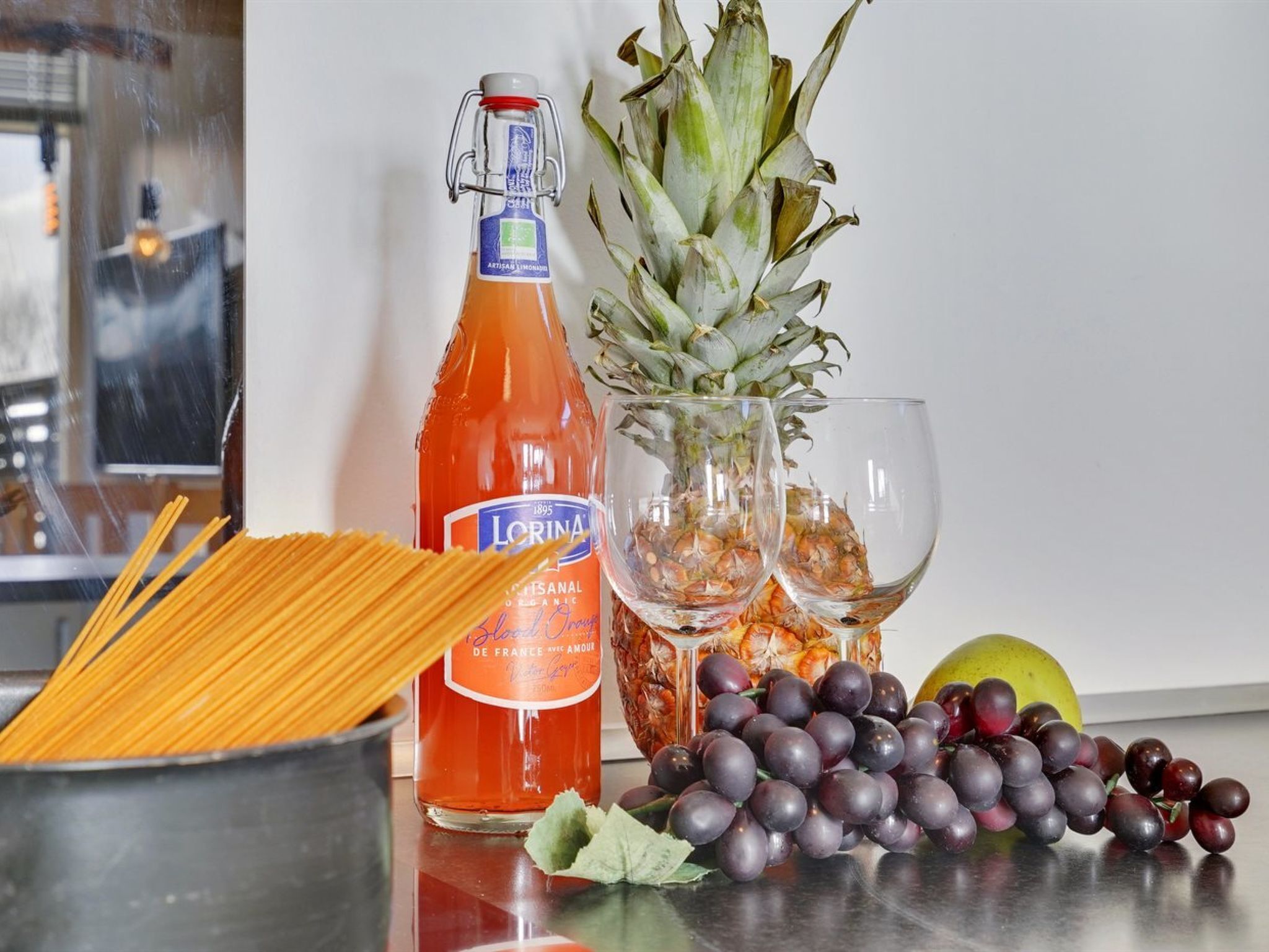 Un plan de travail de cuisine avec une bouteille de LORINA, un ananas, une grappe de raisins, un verre d’eau et un bol de spaghettis.