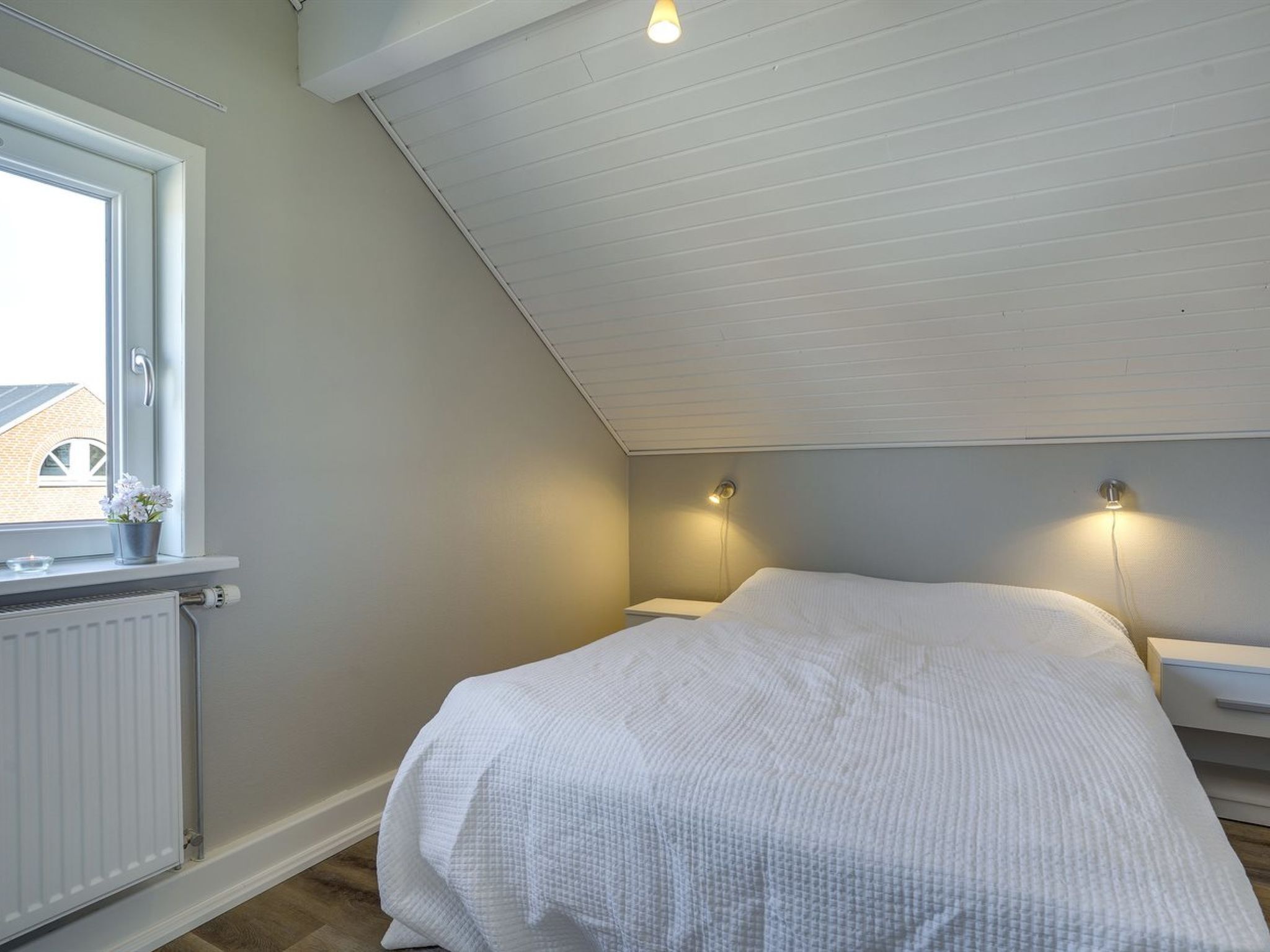 Een slaapkamer met een schuine kap, een raam met uitzicht op een gebouw, een radiator, een bed met een witte spreidek en twee wandlampen.