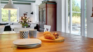 A wooden dining table with plates, cups, and pastries in a bright room with large windows and a green ceiling.