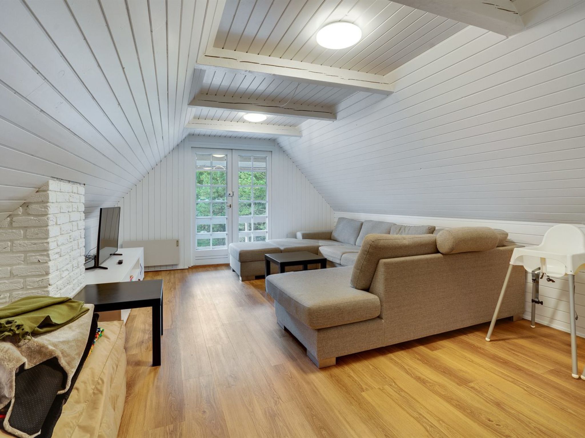 A loft-style room with a sloped ceiling, wooden flooring, a sofa, a small table, and a window with a view of greenery.