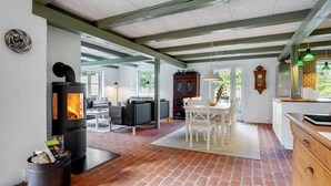 A spacious living area with a brick floor, a fireplace, a dining table with chairs, and a kitchen area with a wooden cabinet and a clock on the wall.