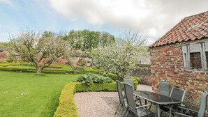 A patio with a table and chairs, a brick building, and a well-maintained garden with flowering trees. - Wolds Way (Driffield)