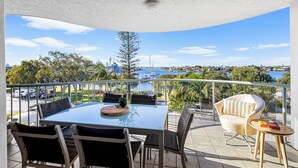 Un balcon avec une table et des chaises en verre, donnant sur une marina où sont amarrés des bateaux, et sur le paysage urbain.