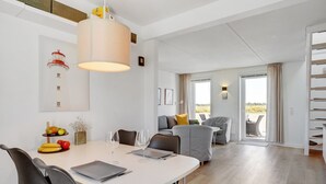 Una sala de estar moderna con comedor, que incluye una mesa blanca, sofás grises y un ventanal con vistas al exterior.