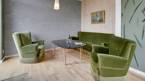 A modern room with a round table, two green armchairs, a green sofa, a wooden wall, and a ceiling light.