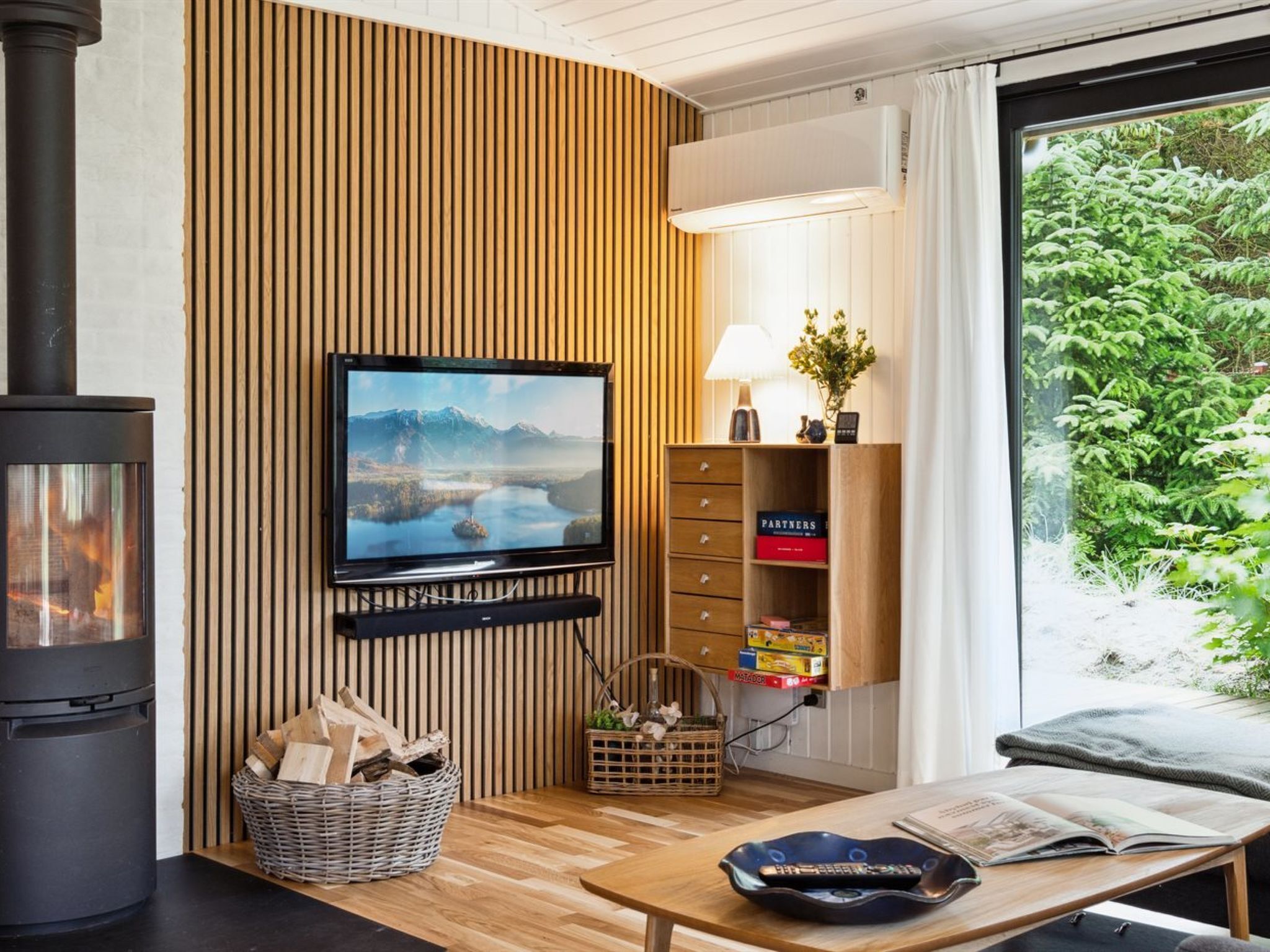 A cozy living room with a fireplace, a mounted TV, a wooden wall panel, a bookshelf, and a window with a view of greenery.