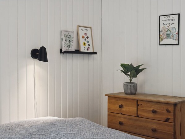 A room with a bed, a wooden dresser, a potted plant, a wall-mounted shelf with books and a framed picture, and a black wall-mounted lamp.
