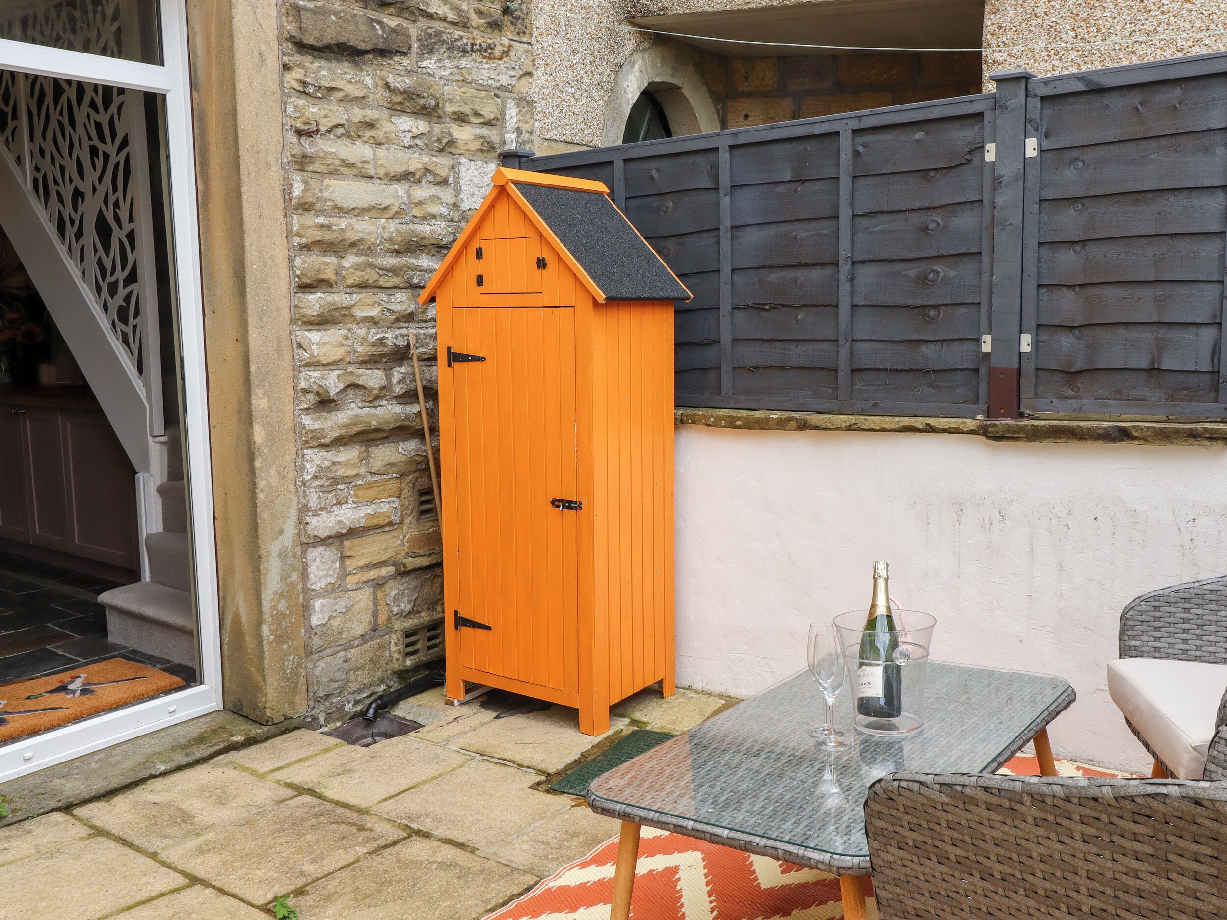 A stone wall, a wooden shed, a glass table with a bottle and glass, and wicker furniture.
