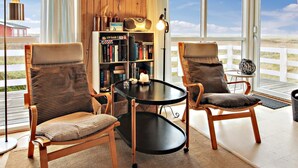Une pièce chaleureuse avec deux fauteuils en osier, une petite table ronde, une bibliothèque et une grande fenêtre donnant sur la plage.