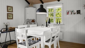 Un coin repas avec une table et des chaises blanches, un luminaire suspendu noir et une fenêtre donnant sur un paysage verdoyant.