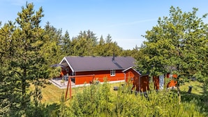 Une cabane rouge au toit en pente, entourée d’une végétation luxuriante et d’arbres.