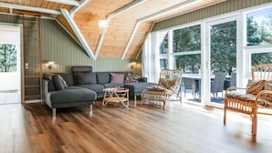 Un loft spacieux avec de hauts plafonds, des poutres en bois, un canapé gris, des fauteuils en osier et de grandes fenêtres donnant sur la verdure.