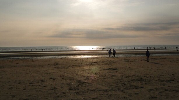 Ein Strand mit tiefstehender Sonne am Horizont, Menschen, die am Ufer entlanggehen, und dem Ozean im Hintergrund.