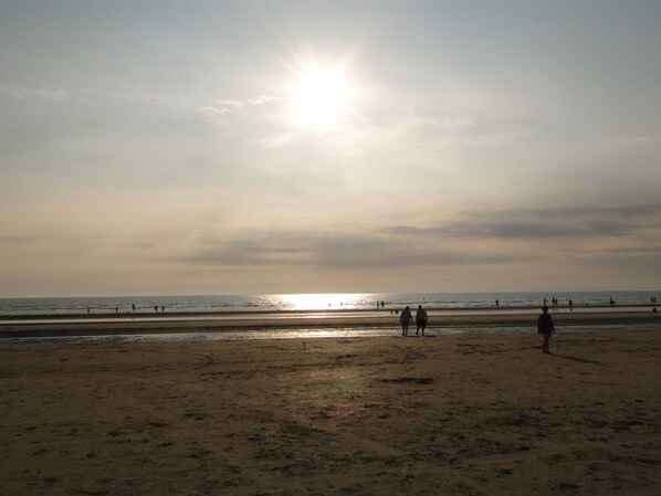 A beach with the sun low on the horizon, people walking along the shore, and the ocean in the background.