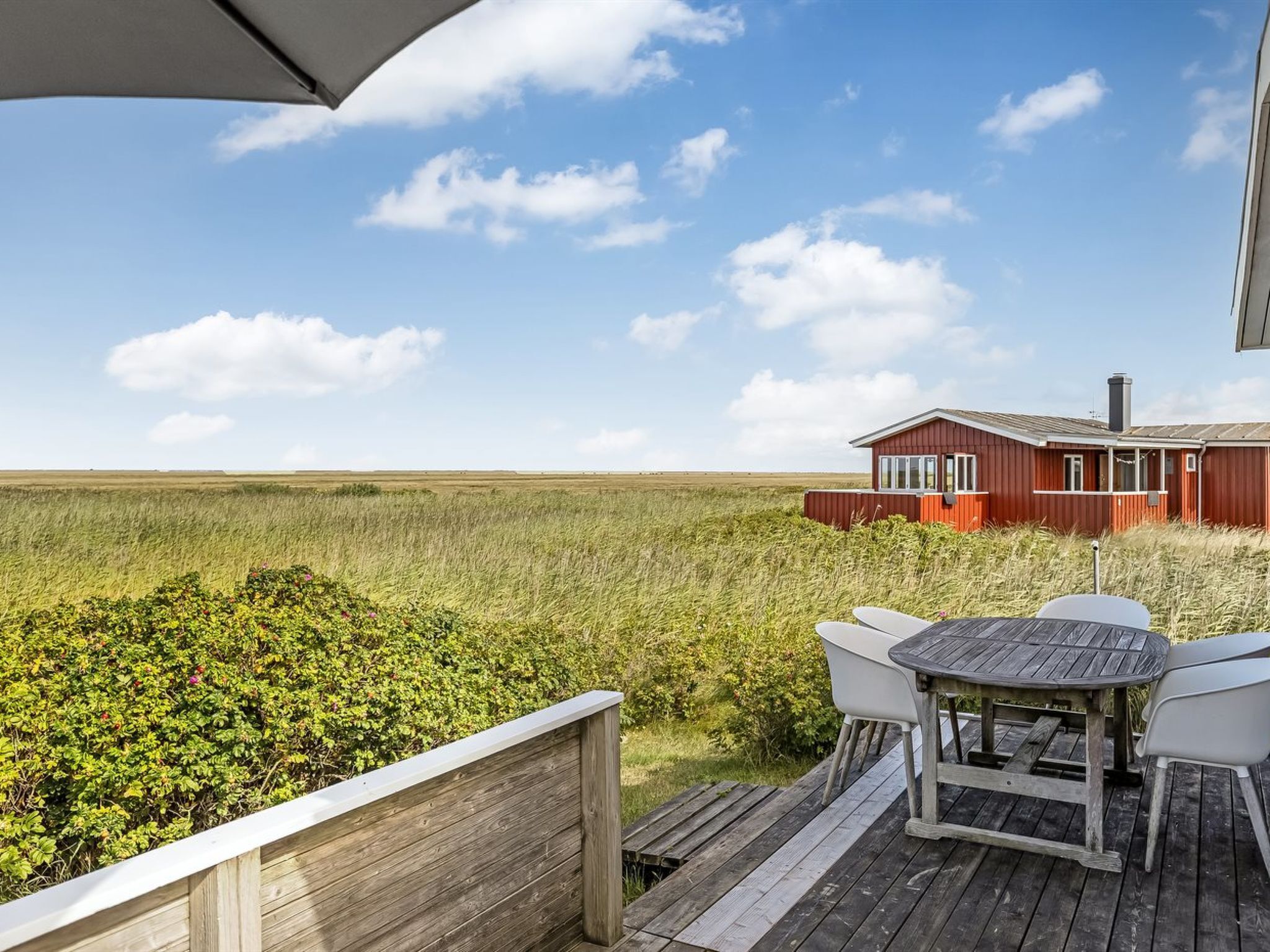 Een houten terras met een tafel en stoelen, met uitzicht op een rood gebouw en een uitgestrekt grasveld onder een heldere lucht.