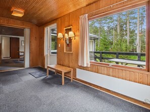 Interior de madeira com um banco, uma janela com vista para árvores e uma porta que dá para outro quarto.