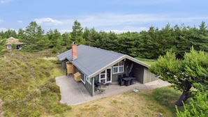 Une petite cabane avec un toit en pente, une cheminée et un espace patio aménagé avec du mobilier d’extérieur.