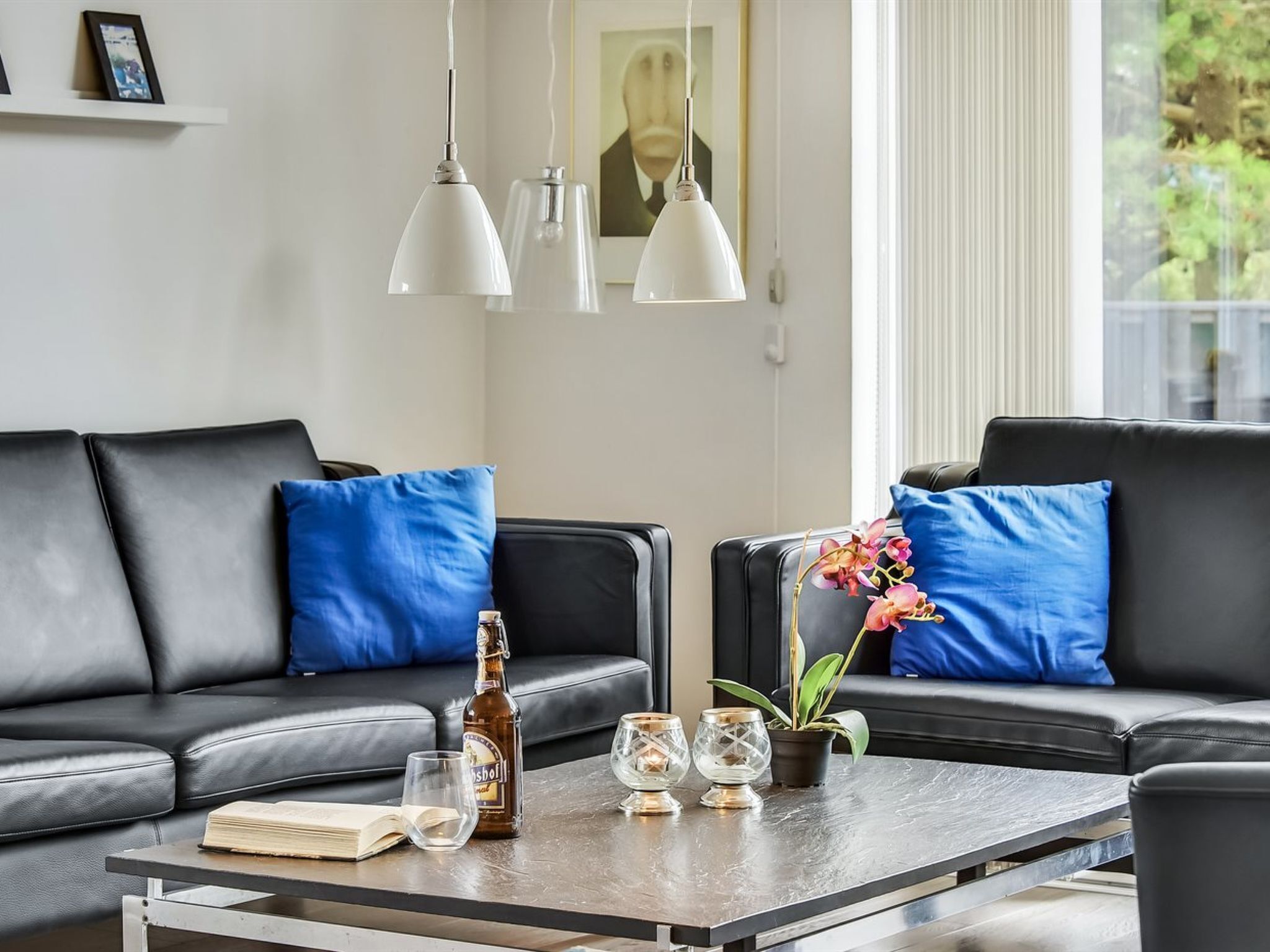 Un salon moderne avec un canapé en cuir noir, des coussins bleus, une table basse sur laquelle se trouvent une bouteille, des verres et un livre.