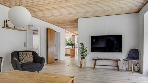 Un salon moderne avec un plafond en bois, un téléviseur fixé au mur, une table à manger en bois et un fauteuil noir.