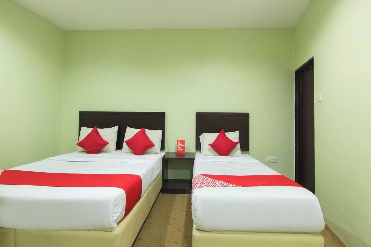 Una habitación con dos camas, cada una con ropa de cama blanca y almohadas rojas, una mesita entre las camas y una puerta a la derecha.