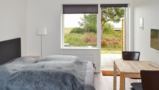 Een slaapkamer met een bed, een tafel, stoelen, een lamp en een raam met uitzicht op groen.