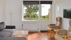 Een moderne woonkamer met een grijze bank, een houten salontafel en twee fauteuils. Een groot raam biedt uitzicht op groen buiten.