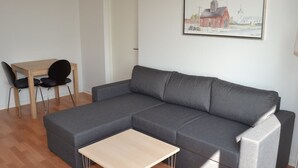 Ett modernt vardagsrum med en grå hörnsoffa, ett soffbord i trä och ett matbord med två stolar. En inramad tavla som föreställer en byggnad hänger på väggen.