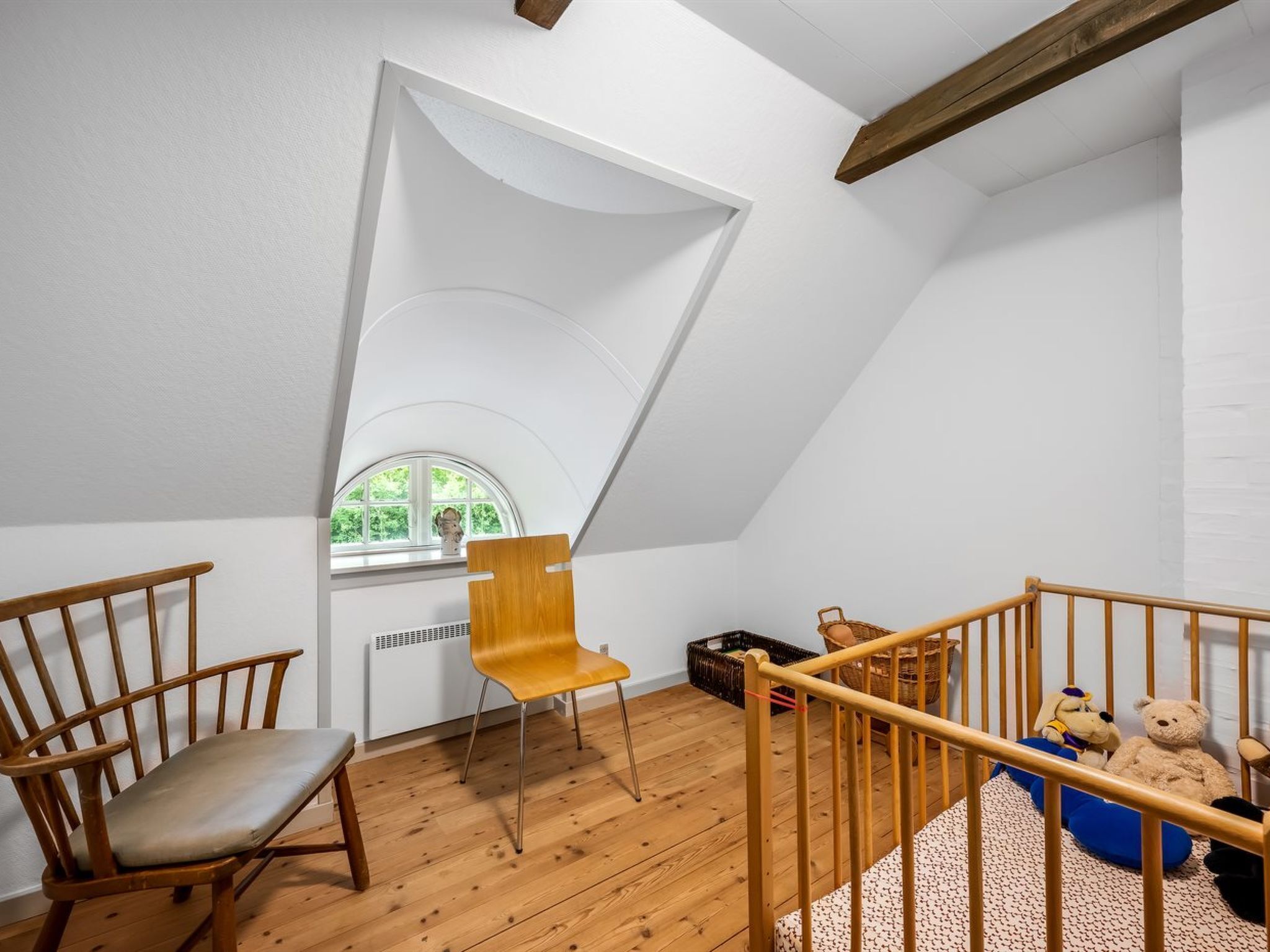 Una habitación con techo inclinado, suelo de madera, una ventana con vistas, una silla de madera, una pequeña mesa y una cuna con peluches.