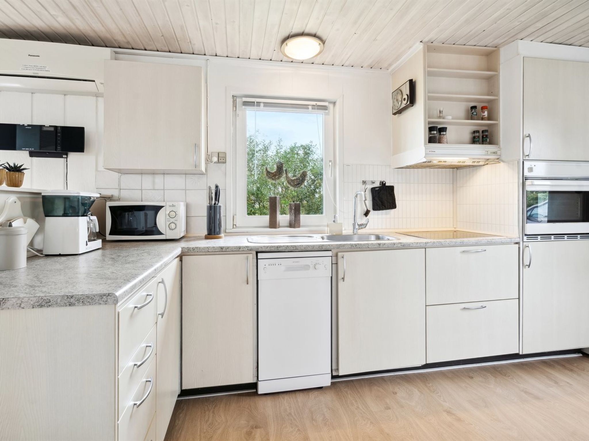 A modern kitchen with white cabinetry, a built-in microwave, a dishwasher, and a window with a view of trees.