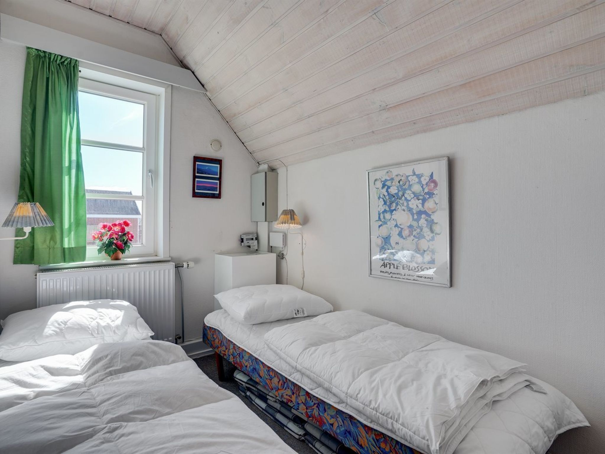Une chambre de dortoir avec deux lits, un radiateur, une fenêtre avec des rideaux verts, un tableau encadré et une lampe.