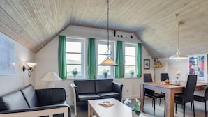 Een gezellige woonkamer met een bank, een salontafel en een eethoek met tafel en stoelen. Er zijn ramen met groene gordijnen en een plafond met houten balken.