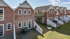 Een rij bakstenen huizen met witte sierlijsten en puntige daken, een klein terras met een tafel en stoelen, en een gazon.