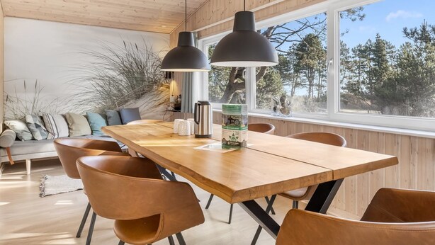 Moderni ruokailutila, jossa on puinen pöytä, nahkaiset tuolit, riippuvalaisimet ja suuri ikkuna, josta avautuu näkymä puita kohti.