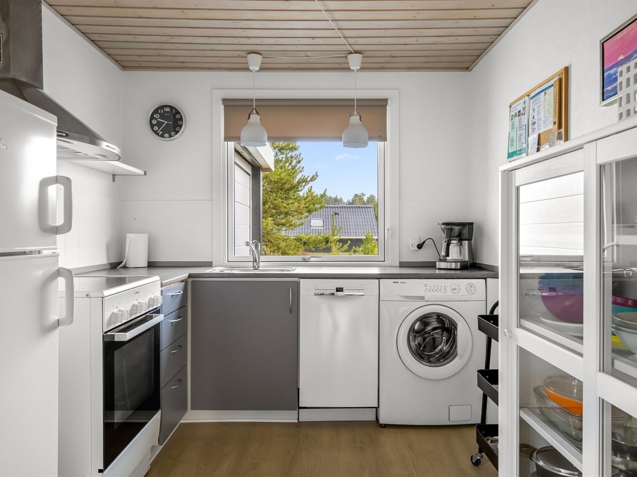 Une cuisine compacte équipée d’un four encastré, d’un lave-linge, d’une machine à café et d’une fenêtre donnant sur un espace verdoyant.