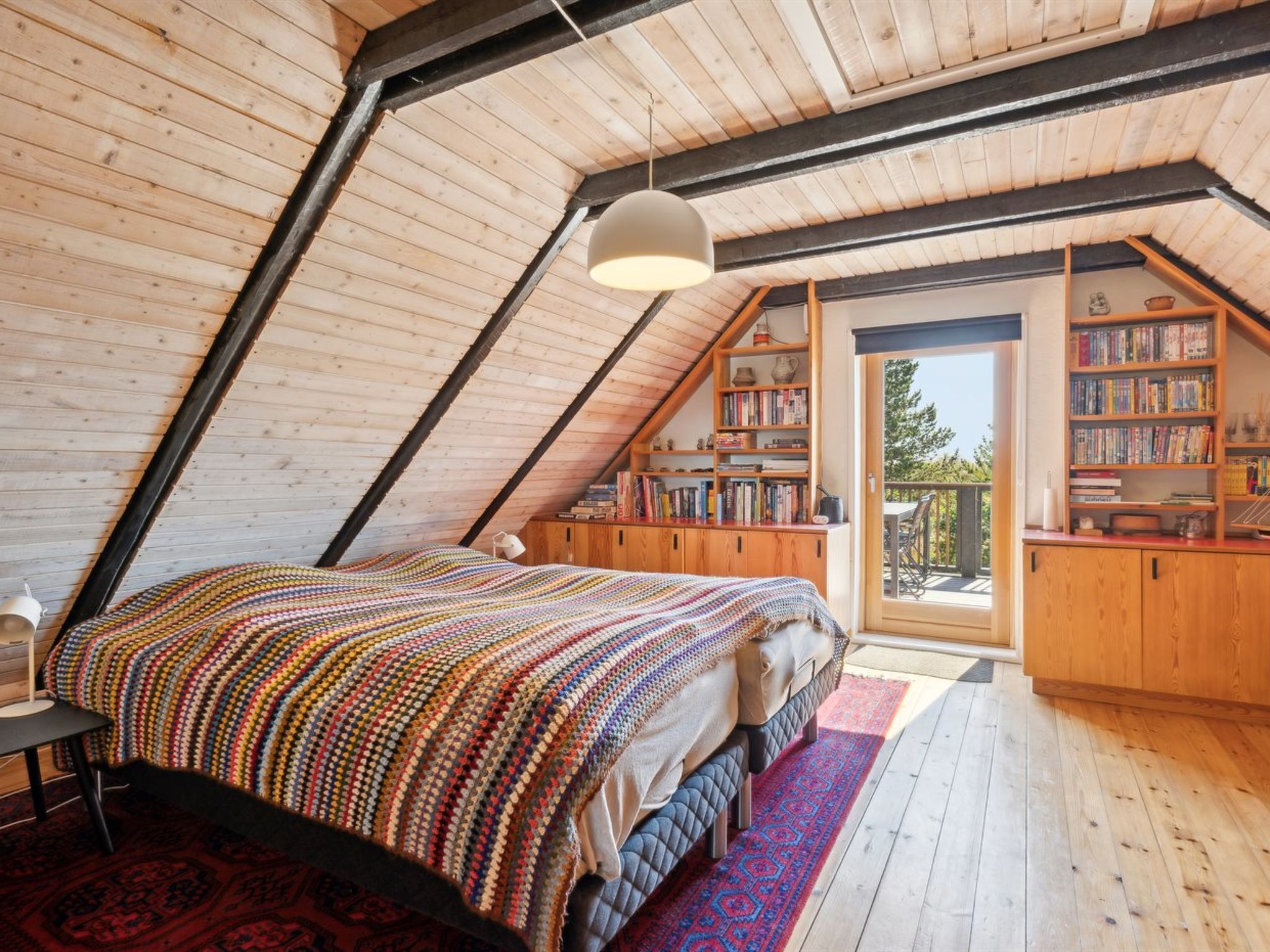 Une chambre mansardée chaleureuse avec un plafond en pente, un grand lit recouvert d’une couverture colorée, une bibliothèque et une vue sur l’extérieur.