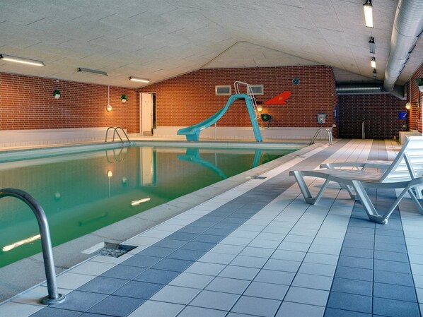 An indoor swimming pool with a blue slide, tiled floor, and lounge chairs.