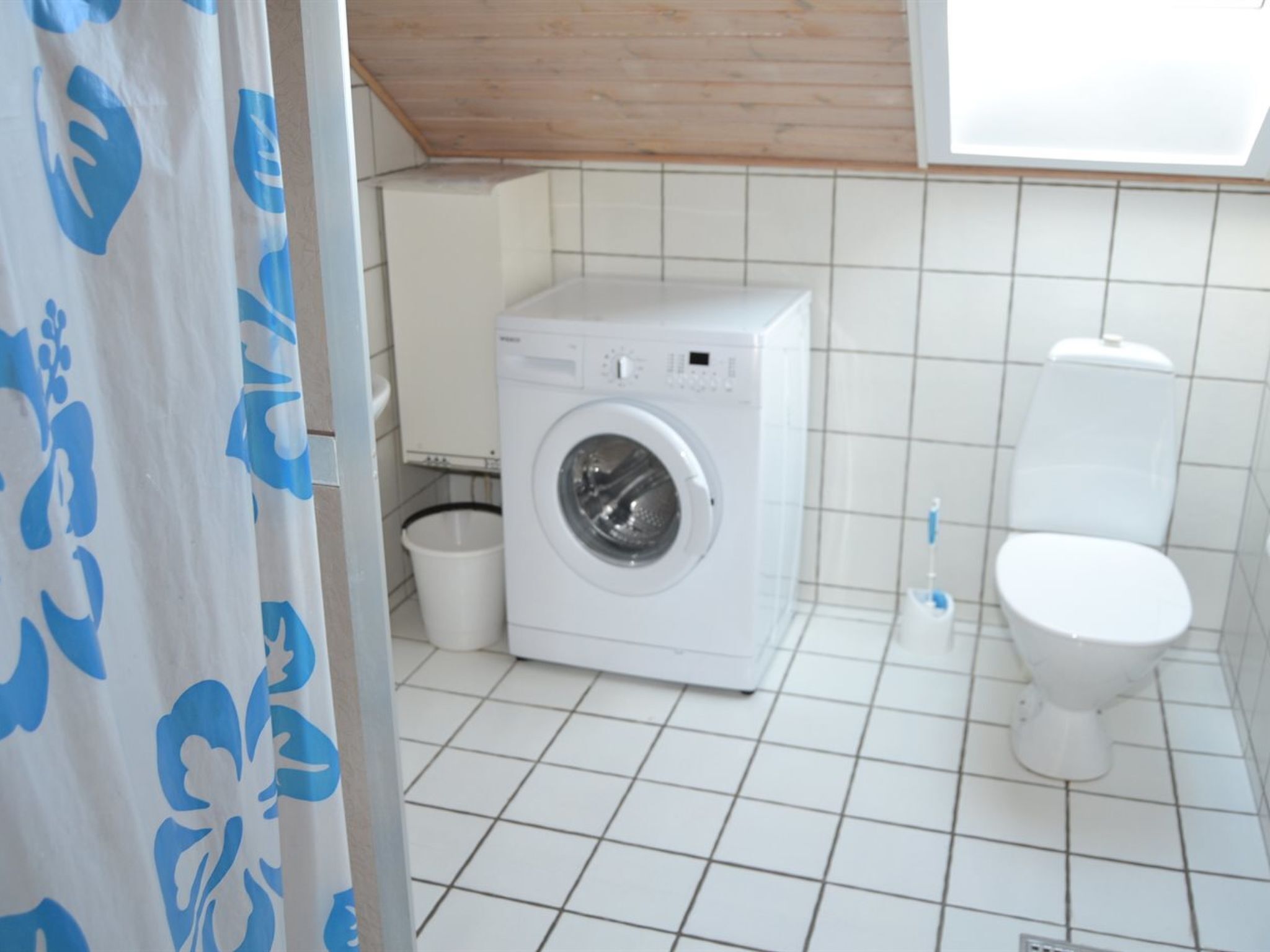 Casa de banho pequena e limpa, com máquina de lavar roupa, sanita e um cesto de lixo.
