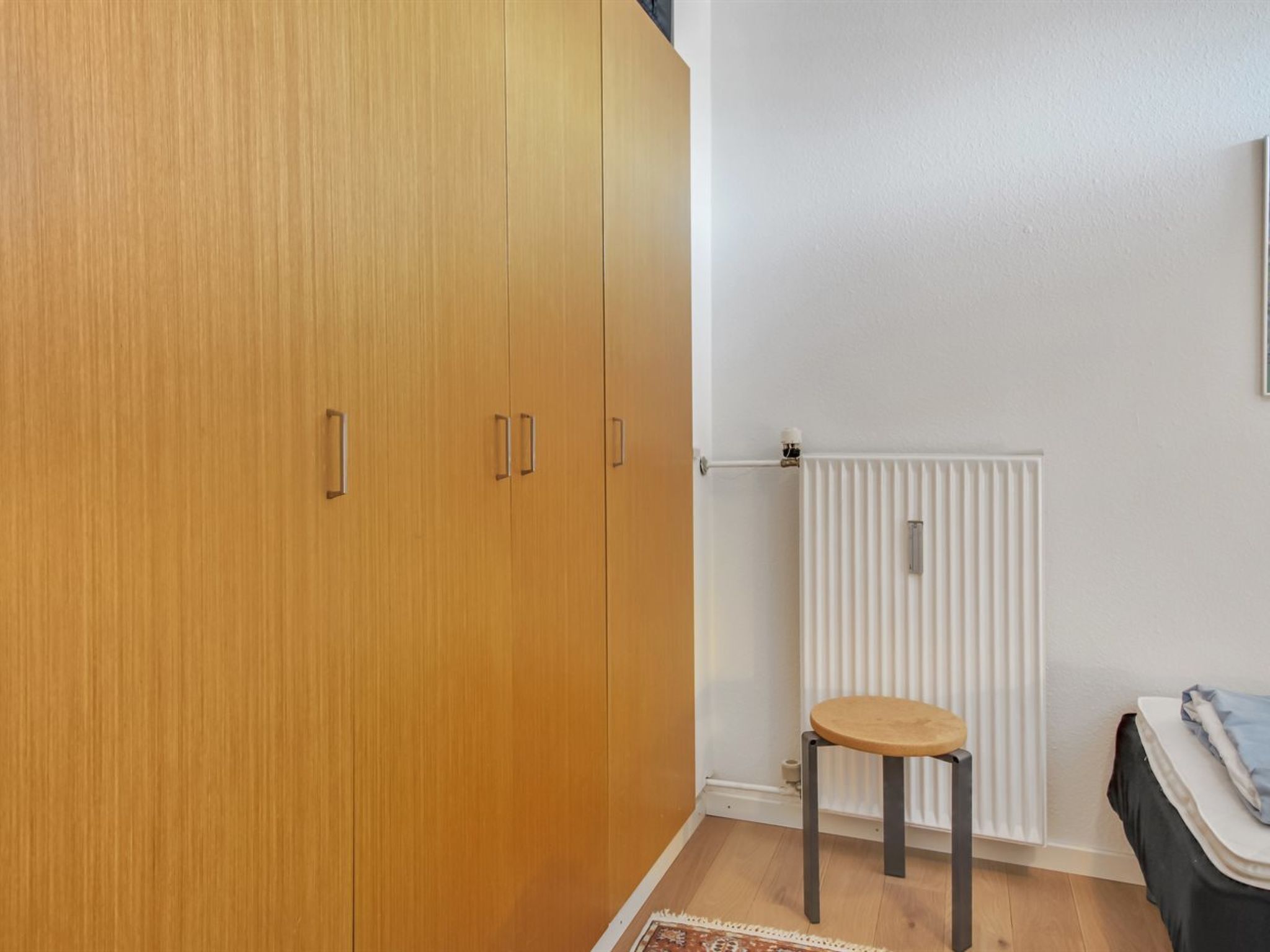 Et værelse med et træskab, en radiator, en lille skammel og en seng med hvid madras og blåt tæppe.