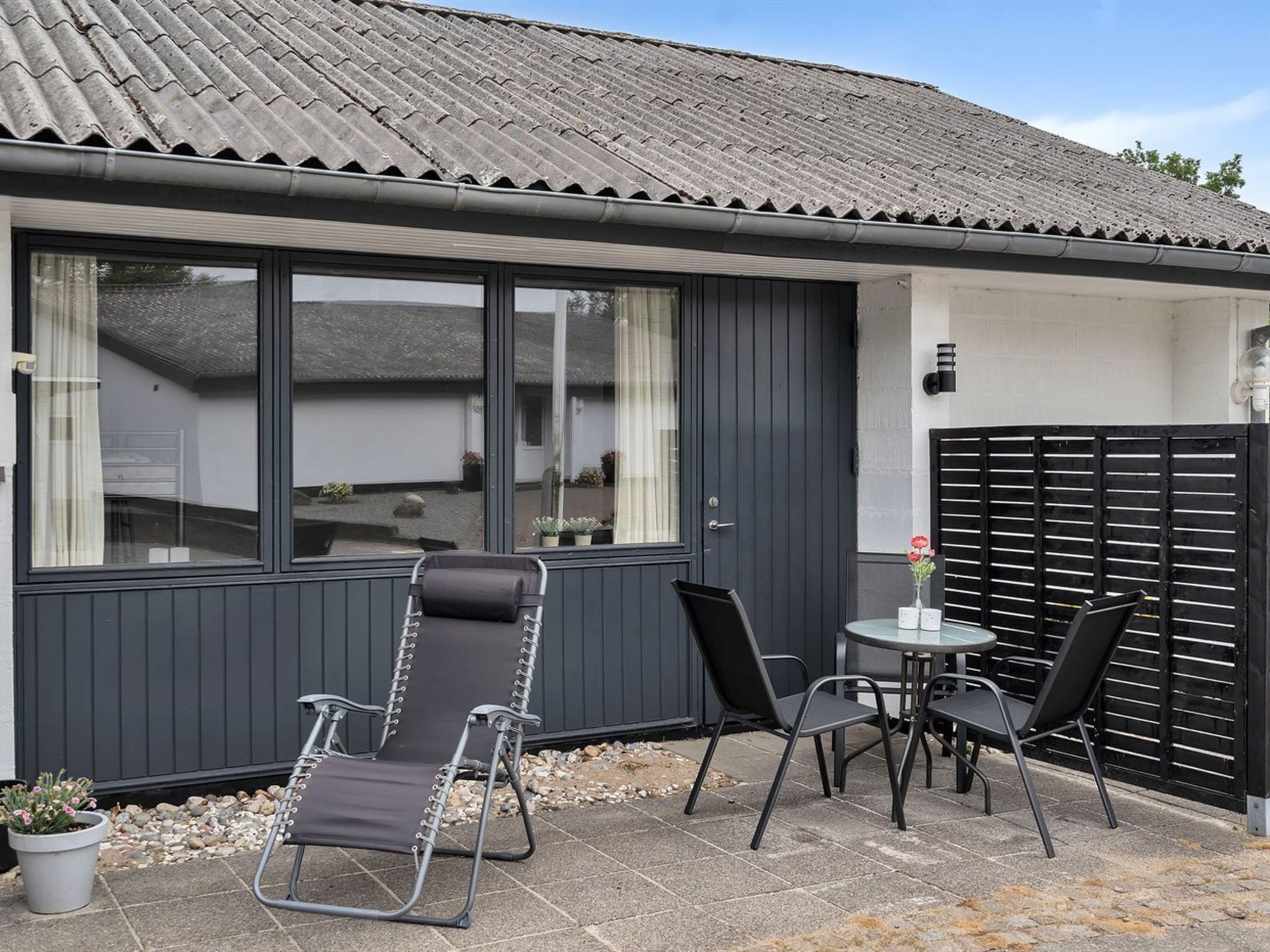 Ein kleiner, moderner Außenbereich zum Sitzen mit Tisch, zwei Stühlen, einem Blumentopf und einem Fenster mit Blick auf das Haus.