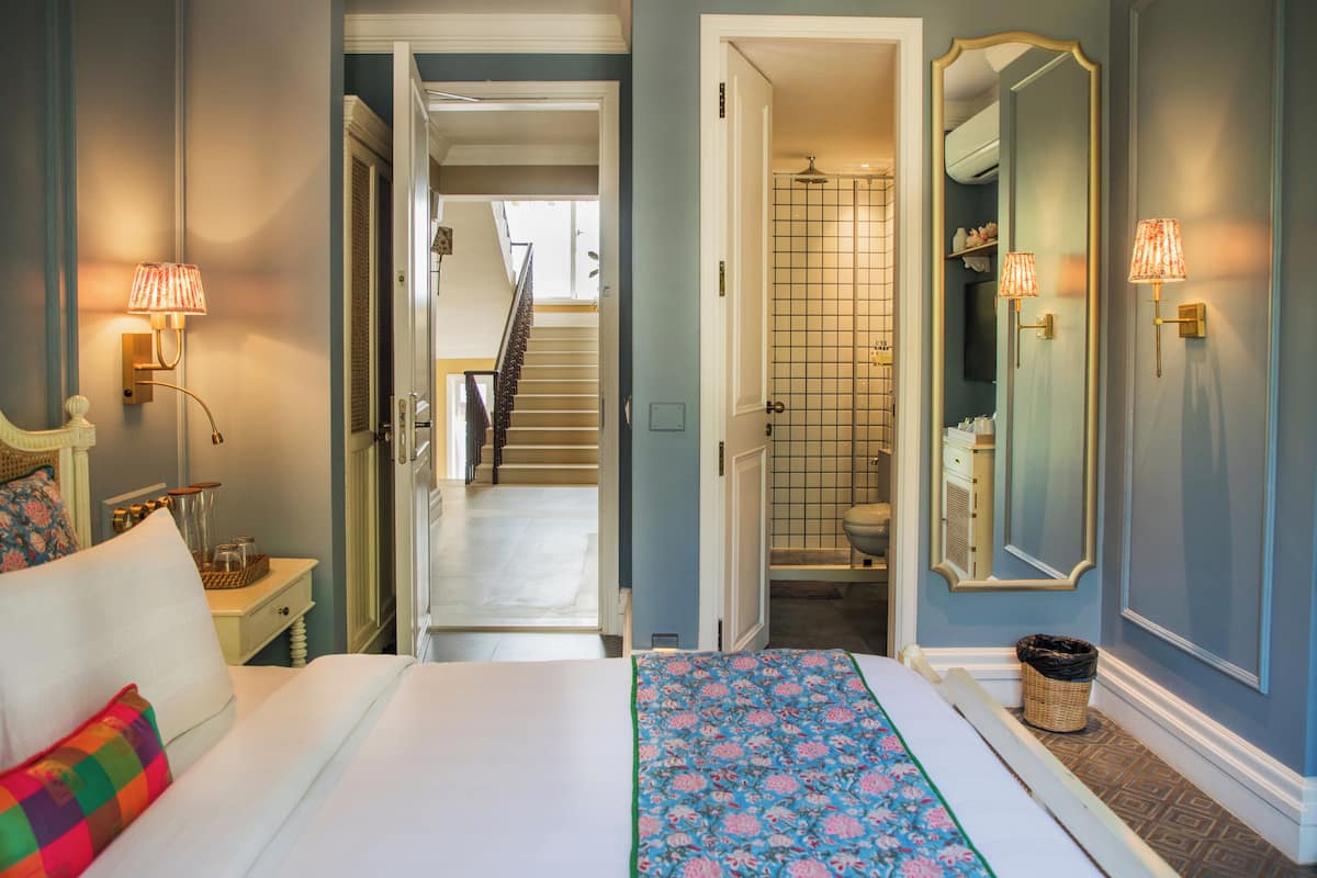 Habitación de hotel con cama, mesita de noche, lámpara, espejo y vistas al baño y la escalera.