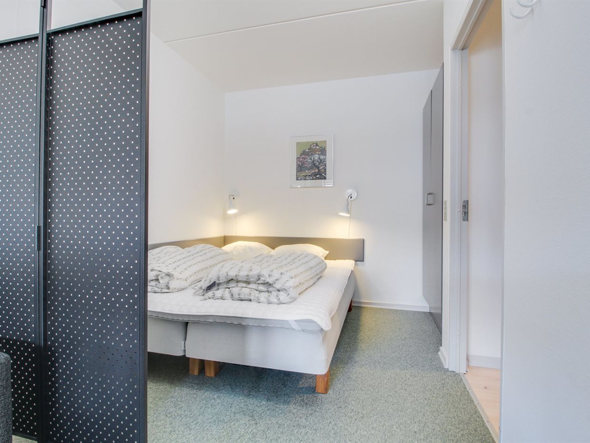 Ein Schlafzimmer mit einem Bett, zwei Nachttischlampen, einem gerahmten Bild an der Wand und einer Tür rechts.