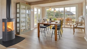 A cozy room with a wood-burning stove, a dining area with chairs, and large windows offering a view of the outdoors. - "Urcu" - 475m from the sea by Interhome (Fanø)