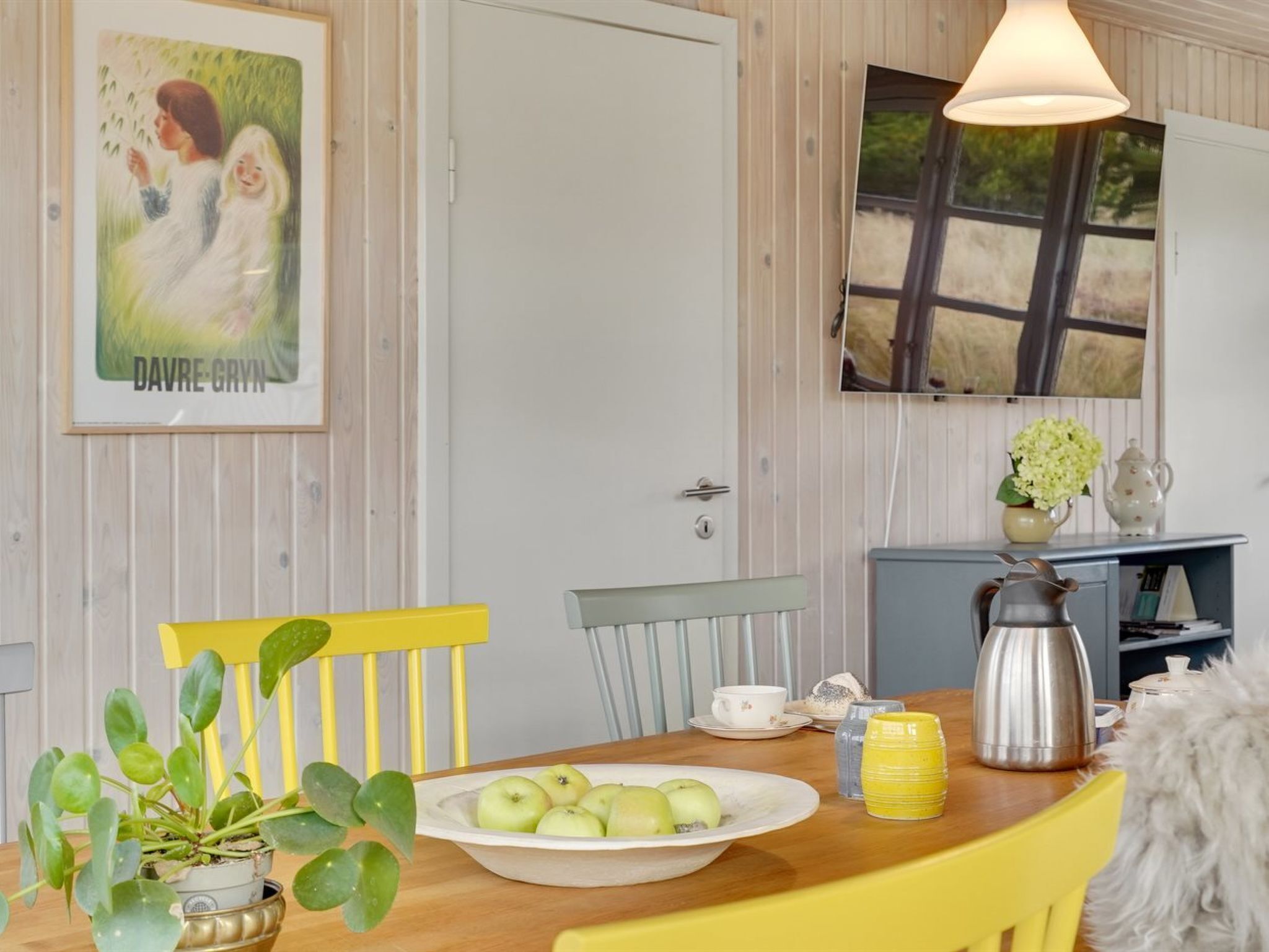 Un coin repas avec une table en bois, des chaises jaunes, un saladier de pommes, une cafetière et une plante en pot.