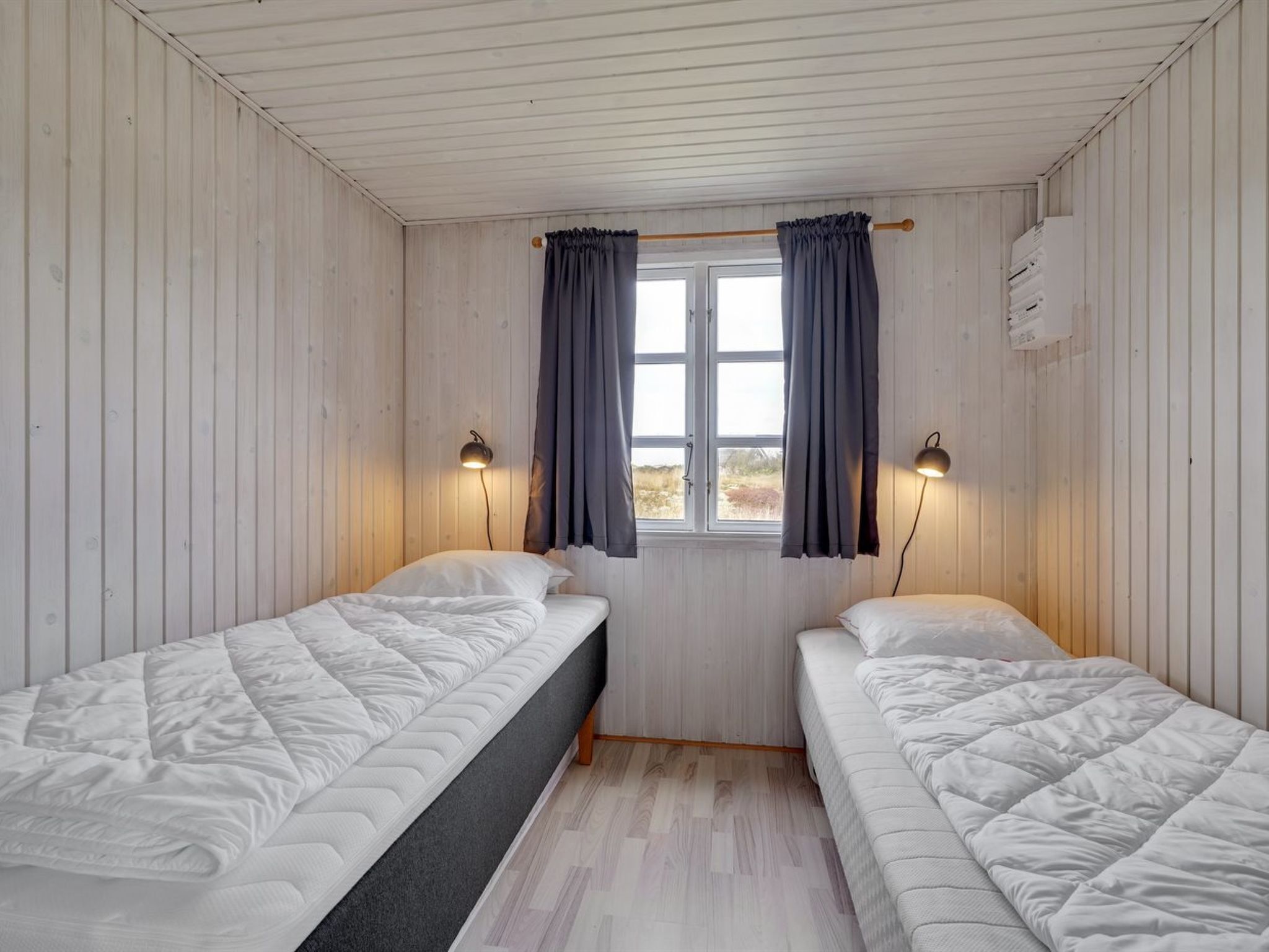 Ein Zimmer mit zwei Betten, Holzwänden, einem Fenster mit Vorhängen und zwei Wandlampen.