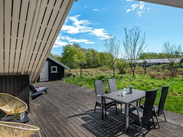 Una terraza de madera con una mesa y sillas, con vistas a un área verde con árboles y un edificio al fondo.