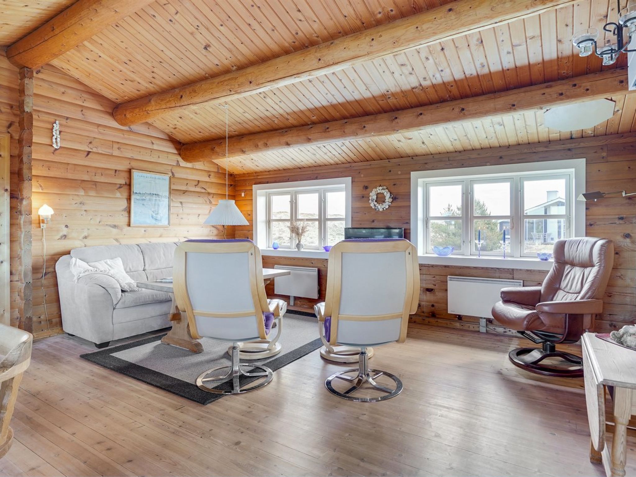 A cozy cabin interior with a wooden ceiling, a sofa, two chairs, a table, and a window with a view.