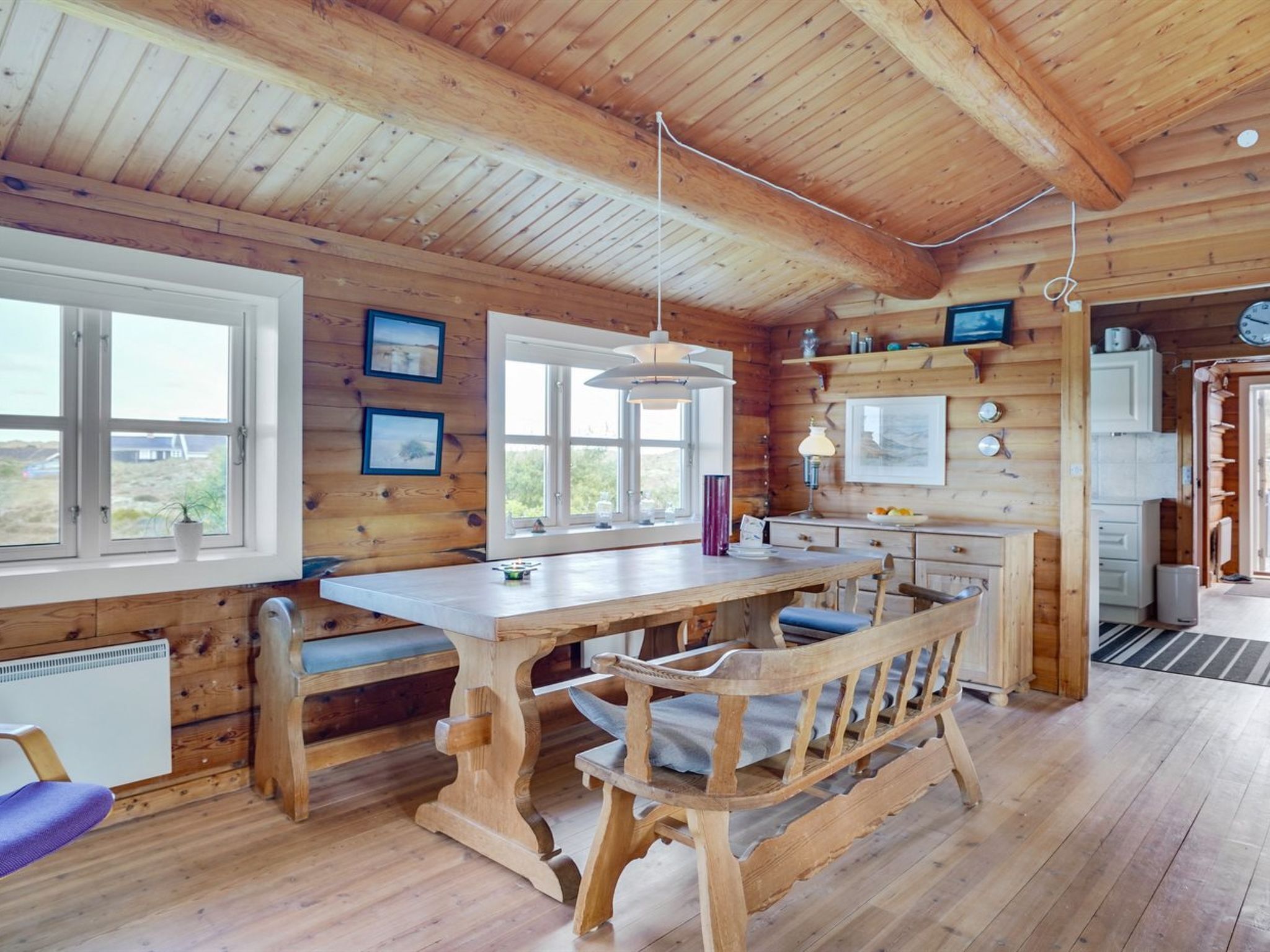 Interior de una cabaña de madera con una mesa de comedor, bancos y una zona de cocina.