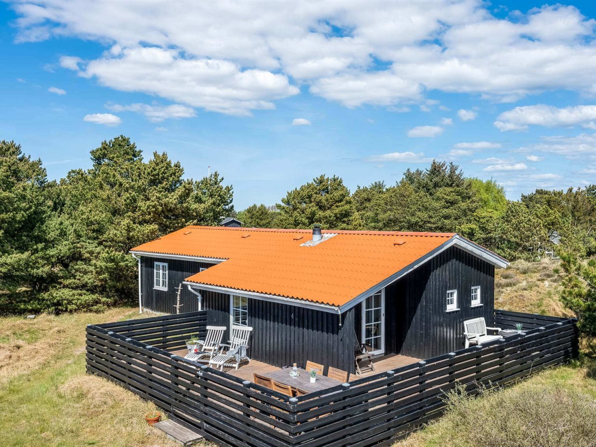 Une cabane à deux étages avec un toit rouge, entourée d’une terrasse en bois et d’une clôture noire.