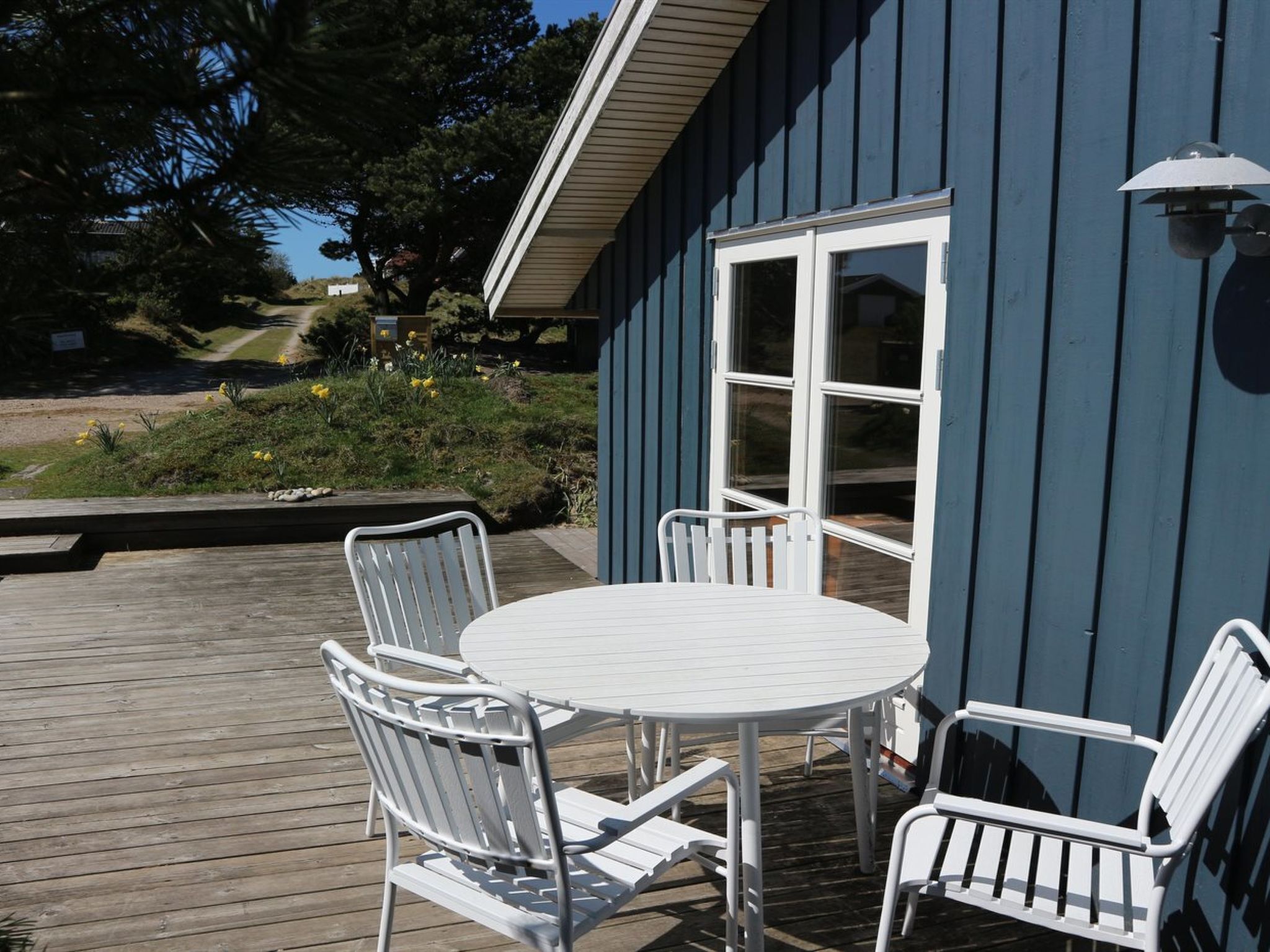 Eine Terrasse mit einem runden Tisch und weißen Metallstühlen, ein blaues Gebäude mit einer weißen Tür und ein Garten mit gelben Blumen.