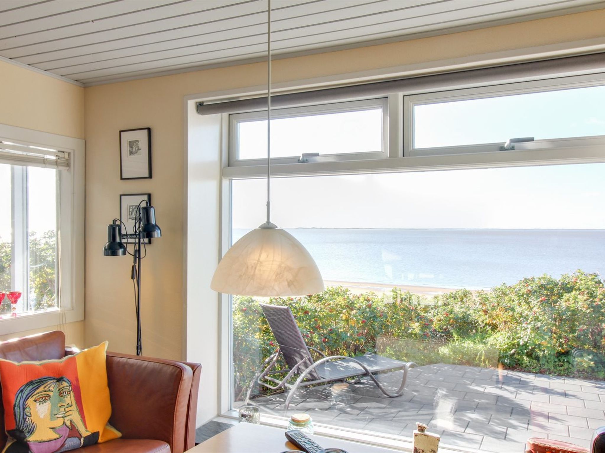 A room with a large window overlooking a beach, a chair, a table with a lamp, and a sofa with colorful cushions.